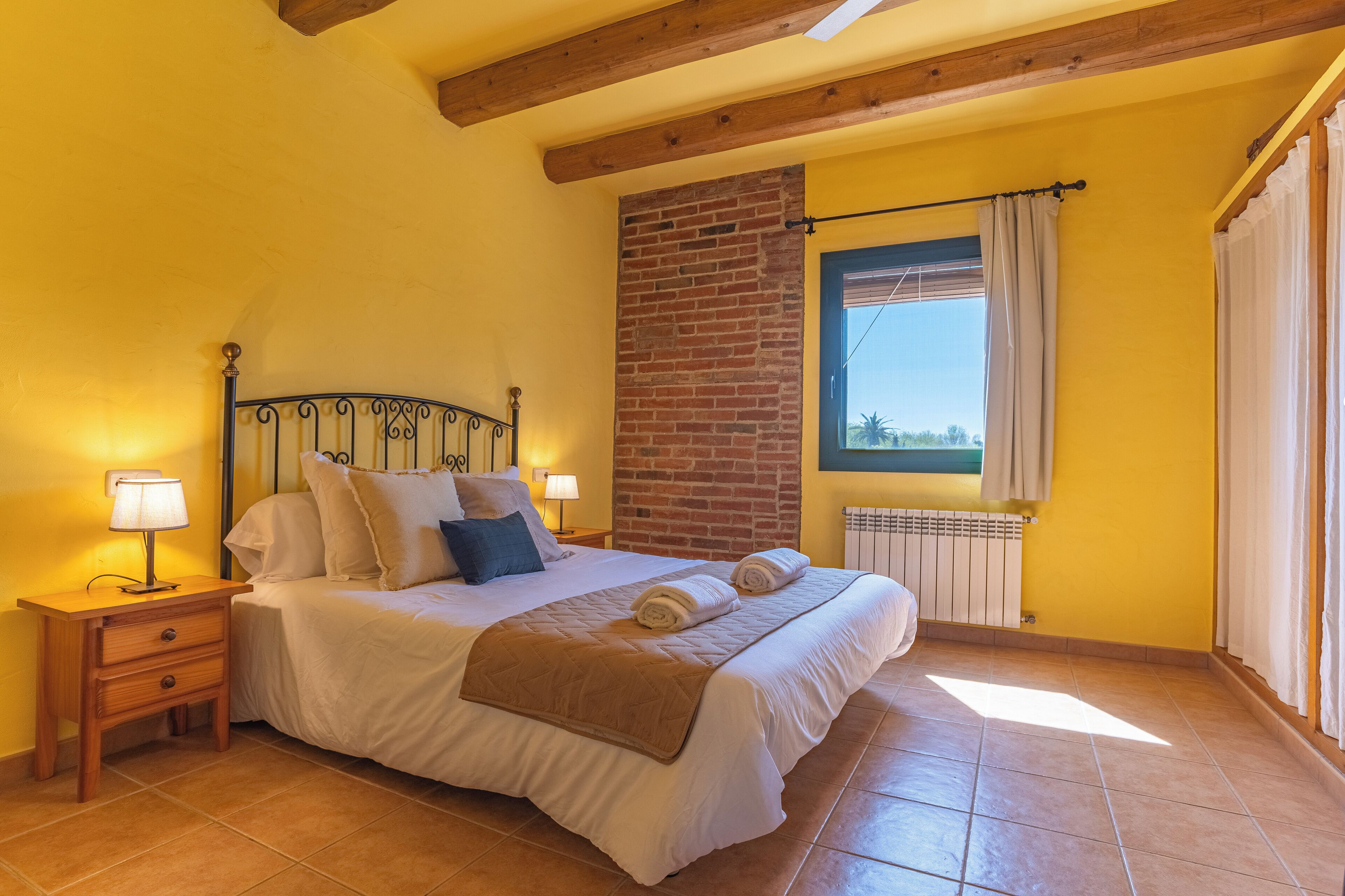 A bedroom with a bed, nightstands, a window with curtains, and a brick wall.