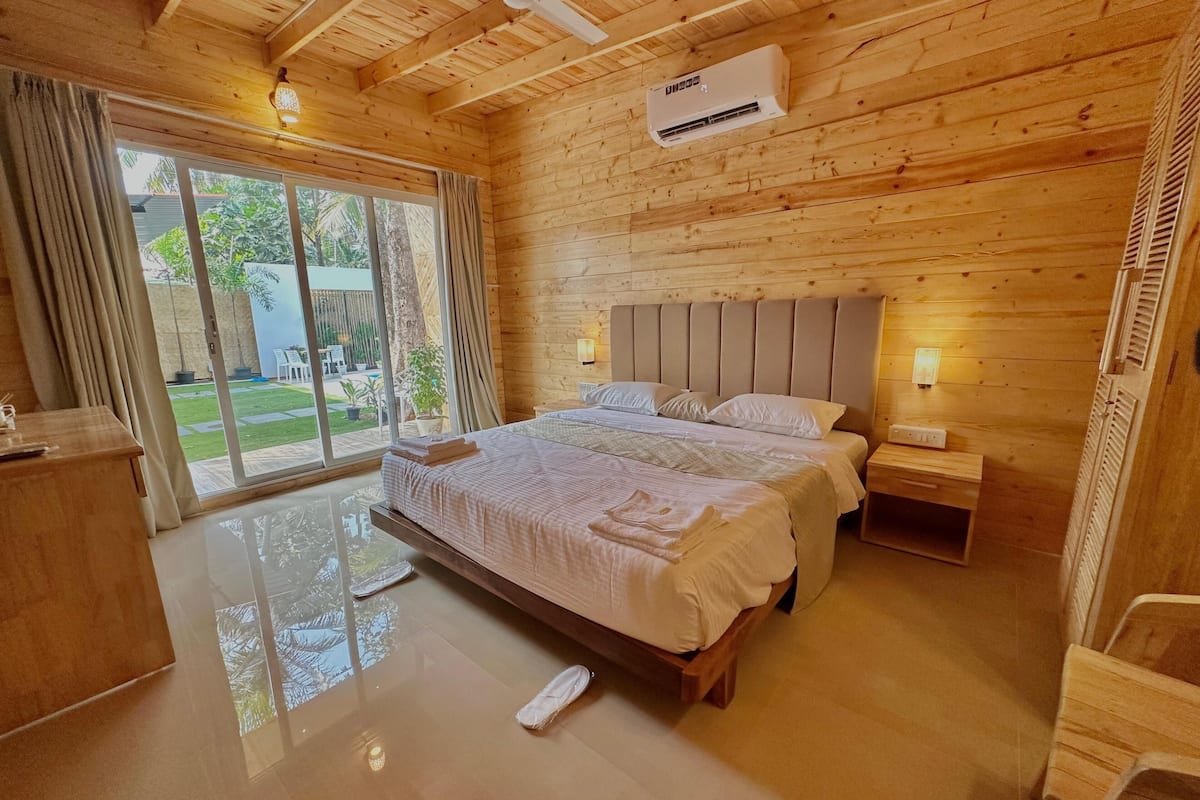 A wooden cabin bedroom with a large bed, a ceiling fan, an air conditioning unit, and a view of the outdoors through a sliding glass door.