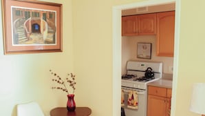 Una cocina con armarios de madera, una estufa y un horno. Se observa una silla blanca y una mesita con un florero.