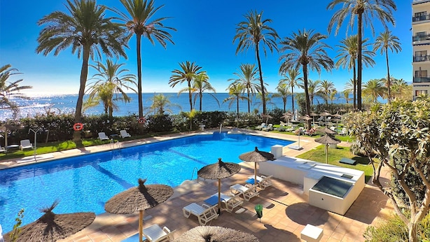 Zona de piscina con sillones de playa y sombrillas, rodeada de palmeras y vistas al mar.