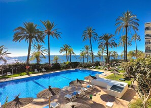 Ein Poolbereich mit Liegestühlen und Sonnenschirmen, umgeben von Palmen und Blick aufs Meer.