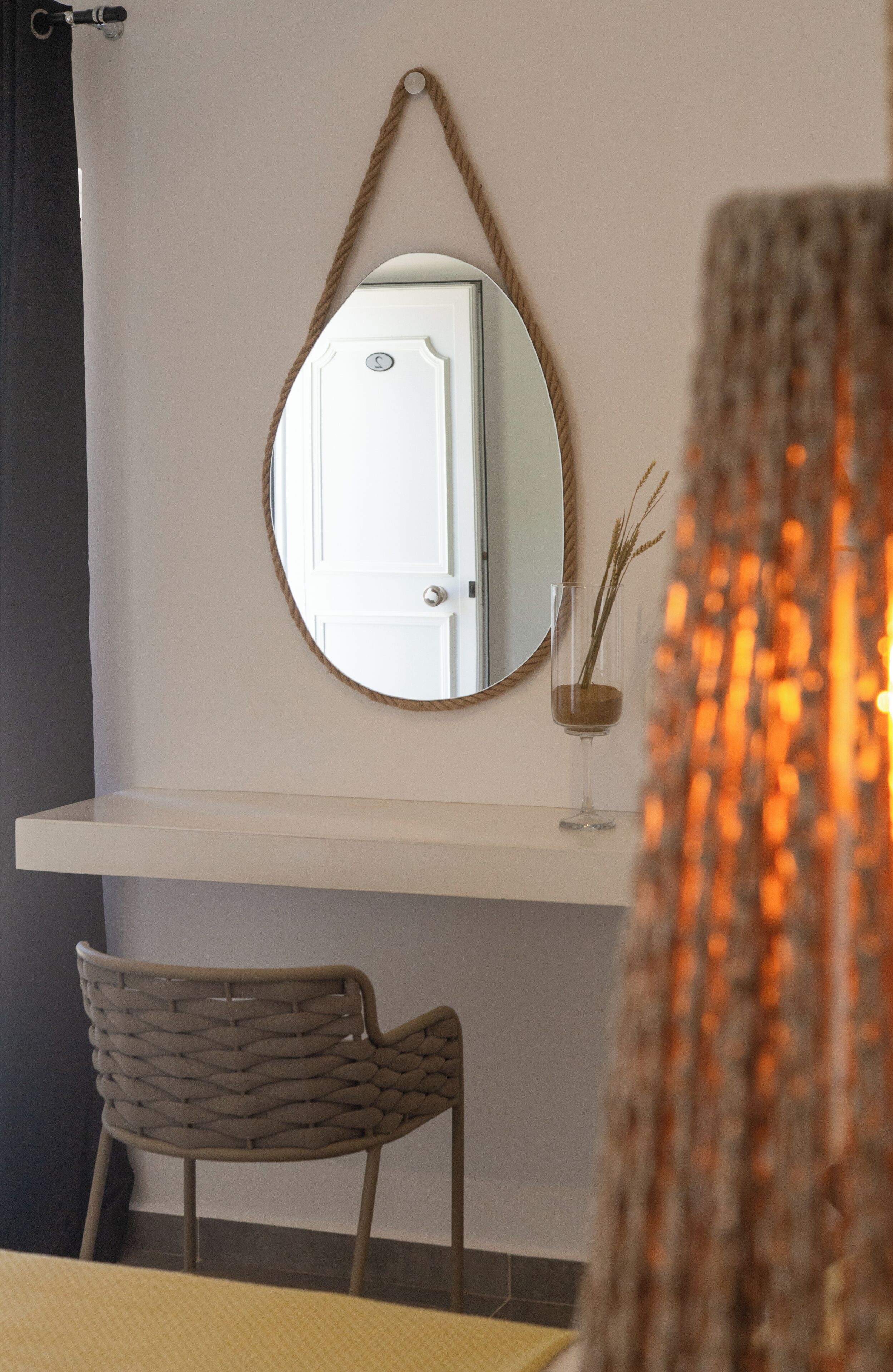 A room with a white wall, a round mirror, a white door, a woven chair, and a glass vase with a plant.