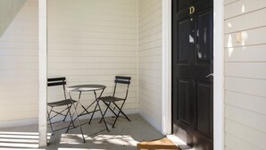A small outdoor seating area with a table and two chairs, a black door with a 'D' marking, and a white exterior wall. - The Moultrie Suite | Walk to Lamar's Sporting Club (Charleston)