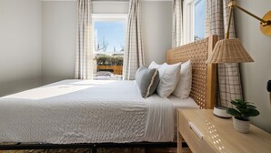 A bedroom with a bed, a bedside table with a lamp, and a view of the outdoors through large windows. - The Moultrie Suite | Walk to Lamar's Sporting Club (Charleston)