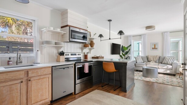 A modern kitchen with white cabinetry, a built-in oven, and a dishwasher. There is a dining area with a gray sofa and a coffee table. - The Moultrie Suite | Walk to Lamar's Sporting Club (Charleston)