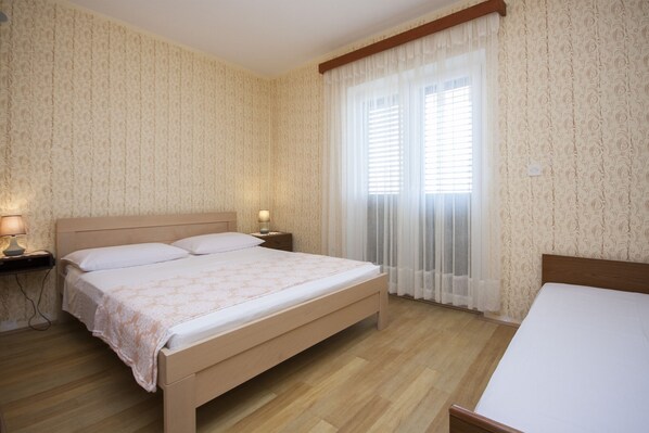 A bedroom with a wooden bed, a nightstand with a lamp, and a window with sheer curtains.