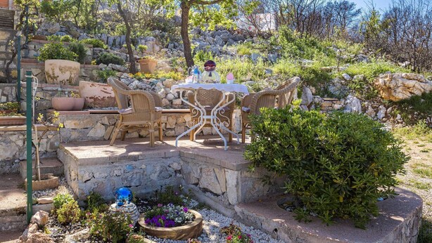 Un espacio exterior con asientos de mimbre y una mesa, rodeado por un jardín con diversas plantas y un cuenco de vidrio decorativo.