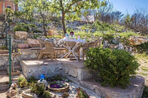 Un espace extérieur aménagé avec des chaises en osier et une table, entouré d’un jardin où poussent différentes plantes et où se trouve un vase décoratif en verre.