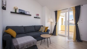 Ein modernes Wohnzimmer mit einem grauen Sofa, einem kleinen runden Tisch und einer Stehlampe. Es gibt einen Balkon mit gelben Vorhängen und Blick ins Freie.