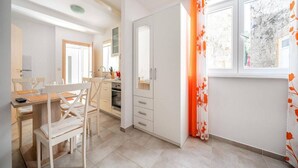 A modern kitchen with white cabinets, a dining area with a wooden table and chairs, and a window with orange curtains. - Studio Apartment Matija Vodice (Vodice)
