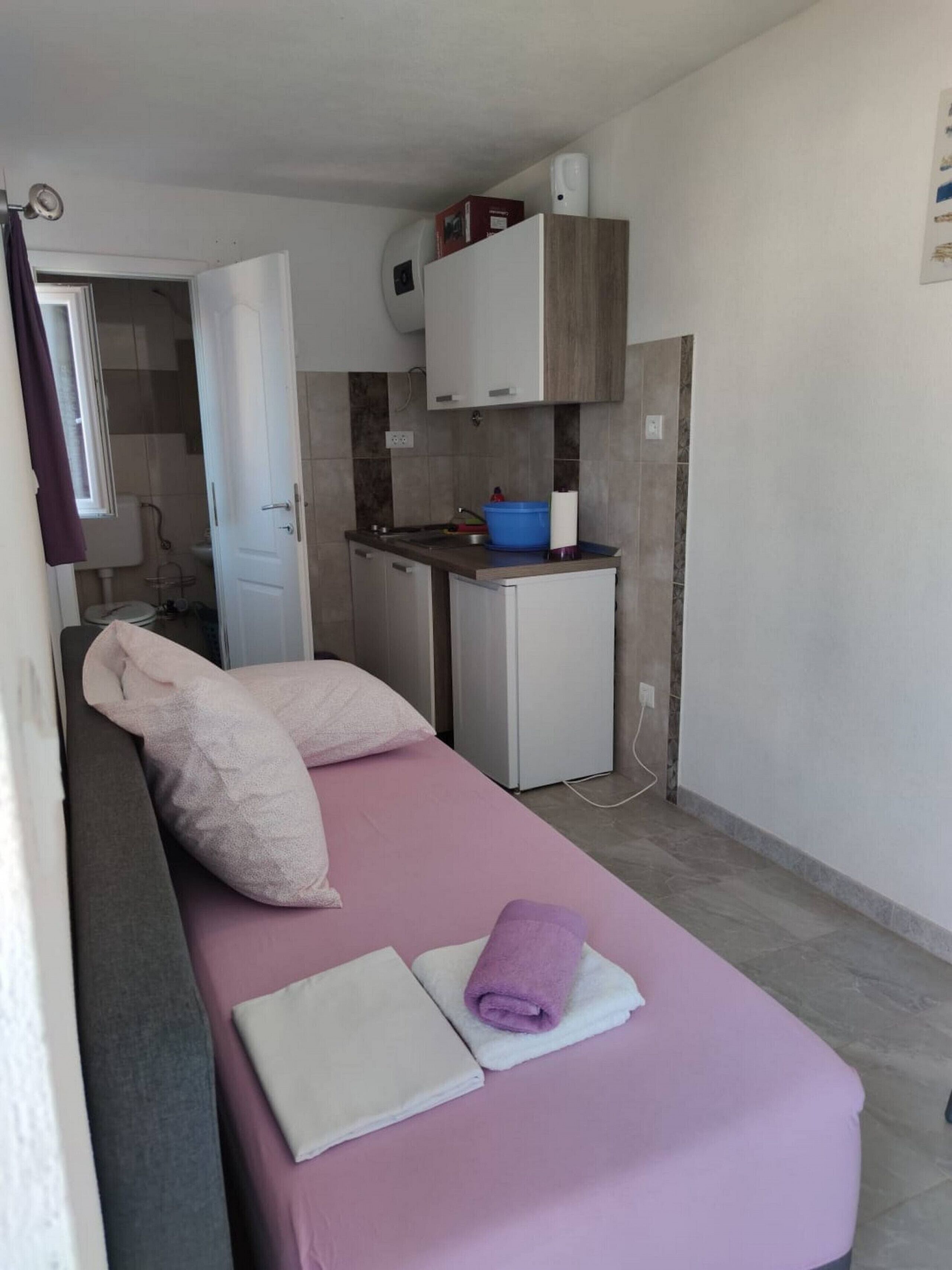 Une petite pièce, de taille modeste, comprenant un lit, un coin cuisine et une salle de bain que l’on aperçoit au fond.