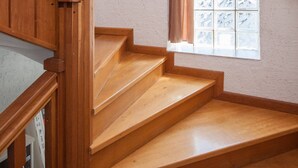 Un escalier en bois, doté d’une rampe, mène à un palier avec une fenêtre et des rideaux.