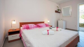 Un lit bien fait, avec une literie rose et blanche, une table de chevet avec une lampe, une petite armoire et une fenêtre avec des stores.