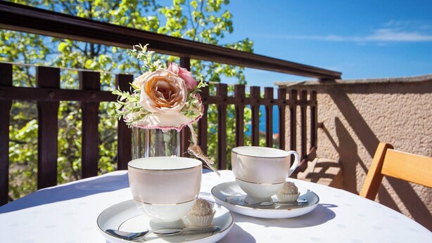 Een tafel met twee kopjes en schotels, een vaas met bloemen en een cupcake op een zonnig terras.