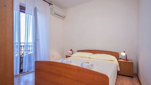 A bedroom with a wooden bed, two bedside lamps, an air conditioning unit, and a balcony with white curtains.