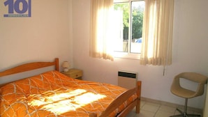 A bedroom with a wooden bed, a bedside table, a chair, a window with curtains, and a radiator. - Large villa 10 pers near beach, 5 bedrooms, quiet (Valras-Plage)