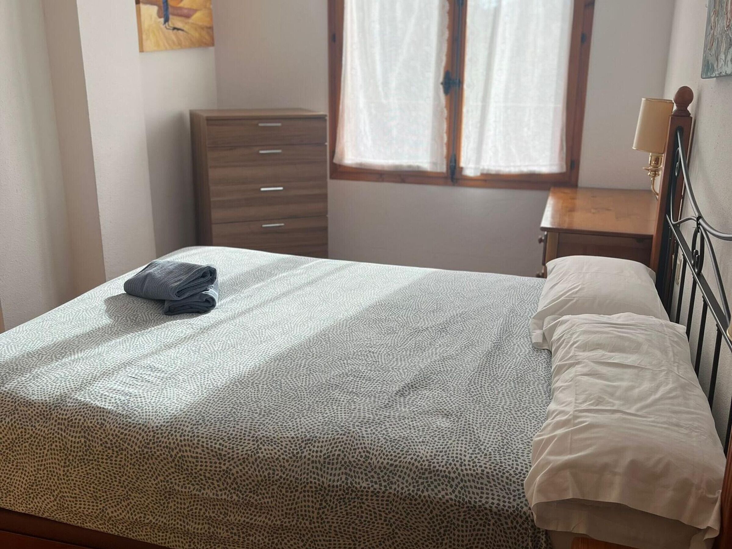 A neatly made bed with a patterned bedspread and a folded towel, a wooden dresser, a bedside table with a lamp, and a window with curtains.
