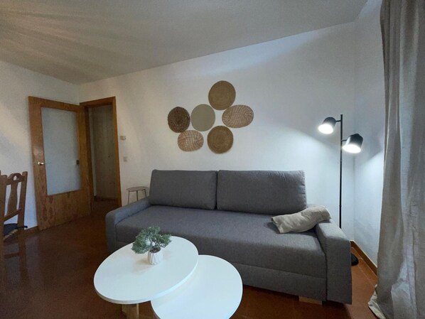 A living room with a grey sofa, a small white table with a plant, and a wall-mounted lamp.
