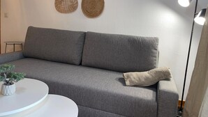 Un canapé gris avec un coussin, une petite table blanche avec une plante, et un mur orné d’une décoration murale tissée.
