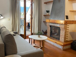 Ein gemütliches Wohnzimmer mit Kamin, einer Couch, einem kleinen Tisch mit einer Pflanze und Blick ins Freie durch eine Schiebetür.