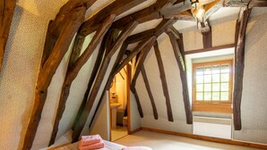 A bedroom with a sloped ceiling, wooden beams, a bed with pink towels, a window, and a bathroom visible through an open door.