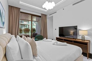 A modern hotel room with a large bed, a flat-screen TV, a bedside table with a lamp, and a view of the city through large windows.