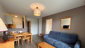 Un salon avec un canapé bleu, une table à manger en bois et des chaises. Une petite table en bois se trouve au centre de la pièce. Une lampe est placée à gauche, et un miroir est accroché au mur.