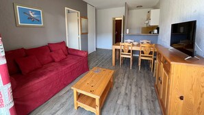 A compact living space with a red sofa, wooden dining table, and chairs, a small wooden coffee table, a TV on a wooden stand, and a kitchen area with a microwave and a sink. - T2 Apartment with terrace and Spassio access, near slopes, 6 people, pet-friendly (Peyragudes)