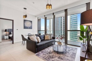 A modern living room with a sofa, coffee table, and dining area. Large windows offer a view of tall buildings. - Luxury 1 Bedroom in the Heart of Famous Downtown (Dubai)