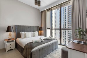 Une chambre d’hôtel moderne avec un grand lit, deux tables de chevet avec des lampes, un bureau avec un ordinateur portable et une grande fenêtre donnant sur la ville.