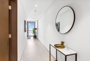 A hallway with a white tiled floor, a wooden door on the left, a round mirror on the right, and a small table with a tray.