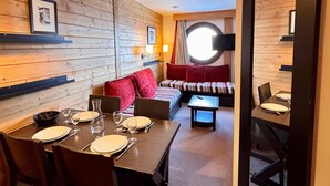 A cozy room with a wooden wall, a round window, a sofa with red cushions, and a dining area with a table set for two.
