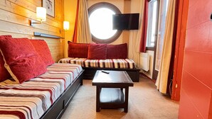 A cozy room with a wooden wall, a round window, a bed with striped bedding, a bench, and a flat-screen TV.