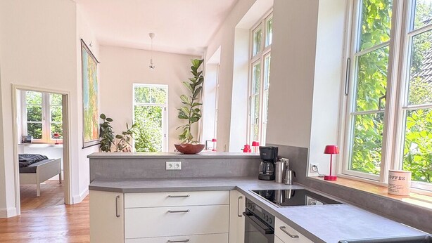 A modern kitchen with white cabinets, a black countertop, and a black rug with 'Kitchen' written on it. There are windows with greenery outside, and a coffee maker on the counter.