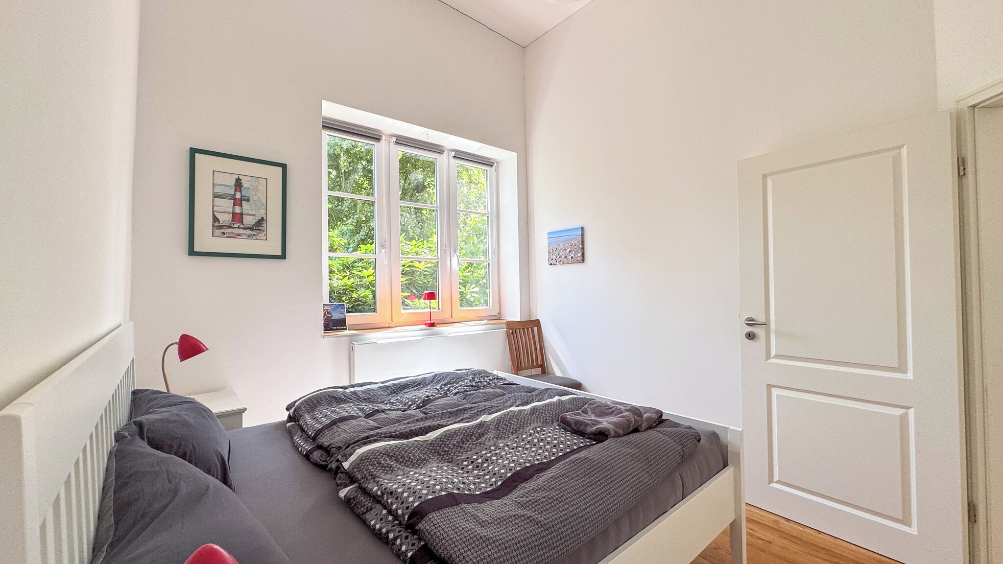 A bedroom with a bed, a window with a view of greenery, a desk, and a door.