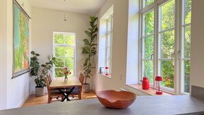 A dining area with a wooden table and chairs, a large window with a view of greenery, and a red lamp on the windowsill.