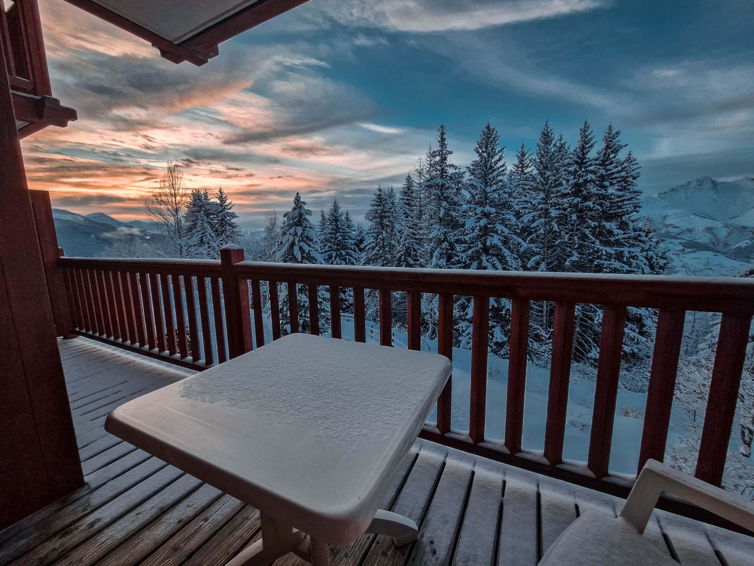 En træterrasse med et bord og stole, med udsigt over et sneklædt bjerglandskab med træer.