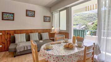 Une pièce chaleureuse avec un banc en bois, une table à manger recouverte d’une nappe à motifs cerisiers, et une vue sur un paysage verdoyant grâce à de grandes fenêtres.