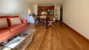 A compact living space with a sofa, a coffee table with a watch and magazine, a wooden dining table with chairs, and a kitchenette in the background.