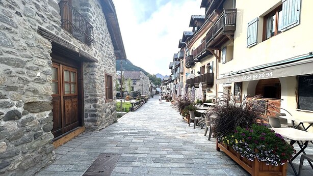 A narrow cobblestone street lined with stone buildings, outdoor seating, and flower planters.