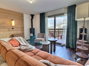 Un salon confortable avec un canapé, un fauteuil et une cheminée. Il y a un balcon avec vue sur l’extérieur.