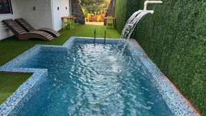 A rectangular swimming pool with a mosaic tile border, a waterfall feature, and a green artificial lawn.