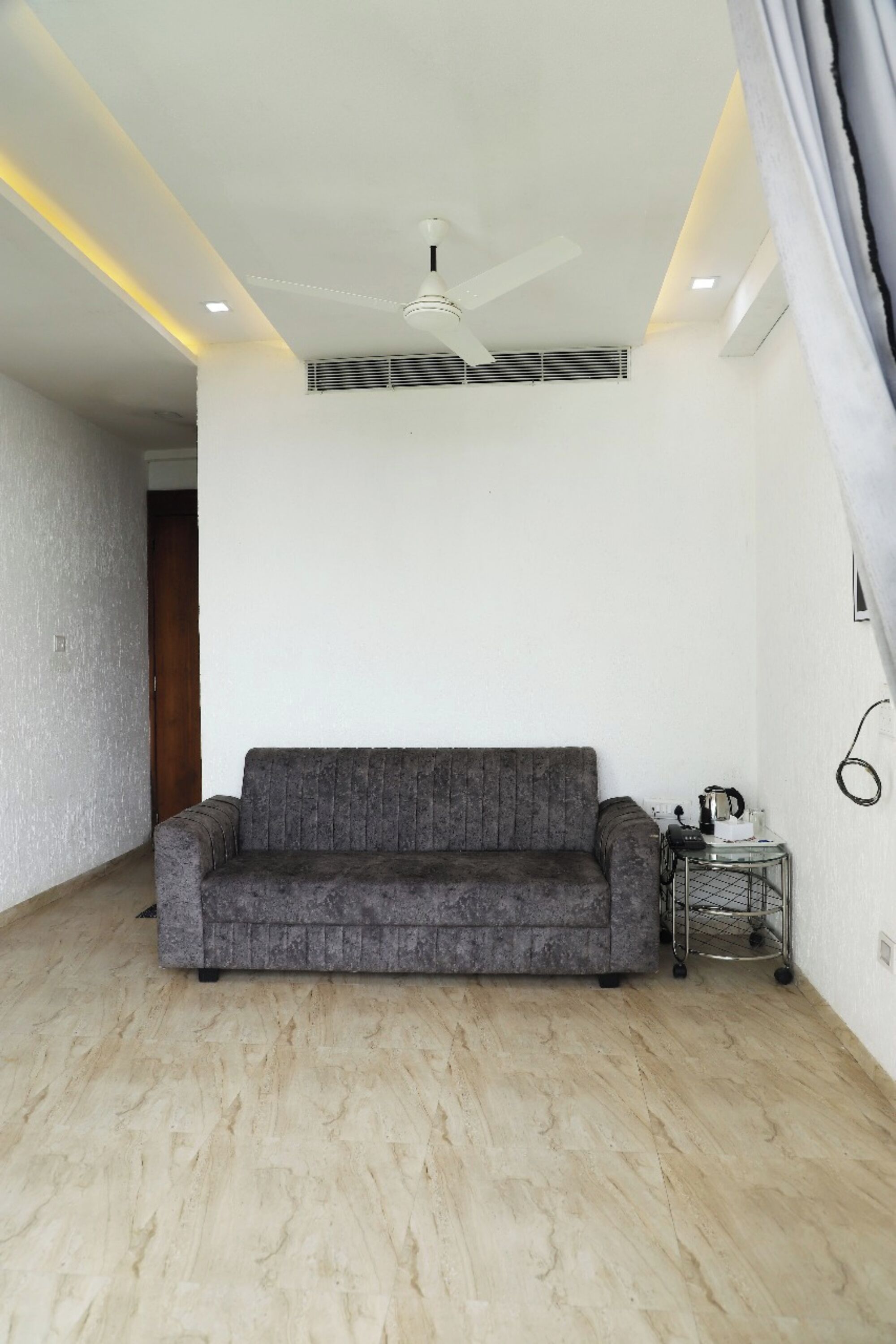 Une pièce avec un canapé gris, une petite table d’appoint sur laquelle se trouve une bouilloire, et un ventilateur de plafond.