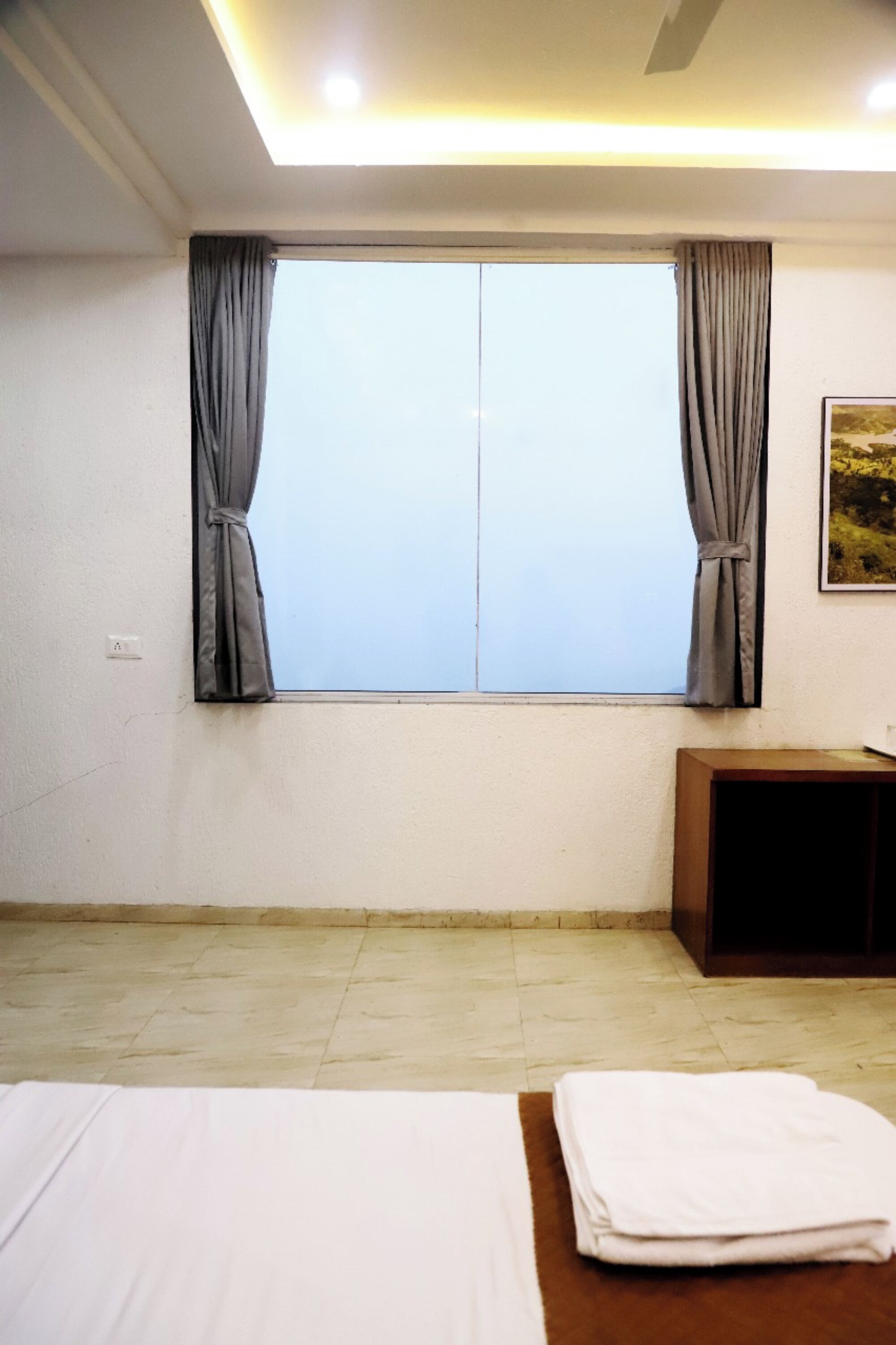 A hotel room with a large window, a bed, a bedside table, and a wall-mounted painting.