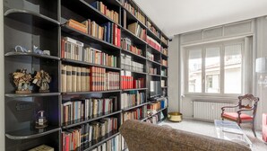 Ein Zimmer mit einer großen Bücherwand, einem Holzstuhl und einem Fenster mit Blick auf Gebäude.