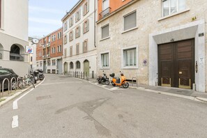 Une rue où sont garés des vélos et des scooters, avec une voiture et un immeuble doté d’une entrée à double porte.