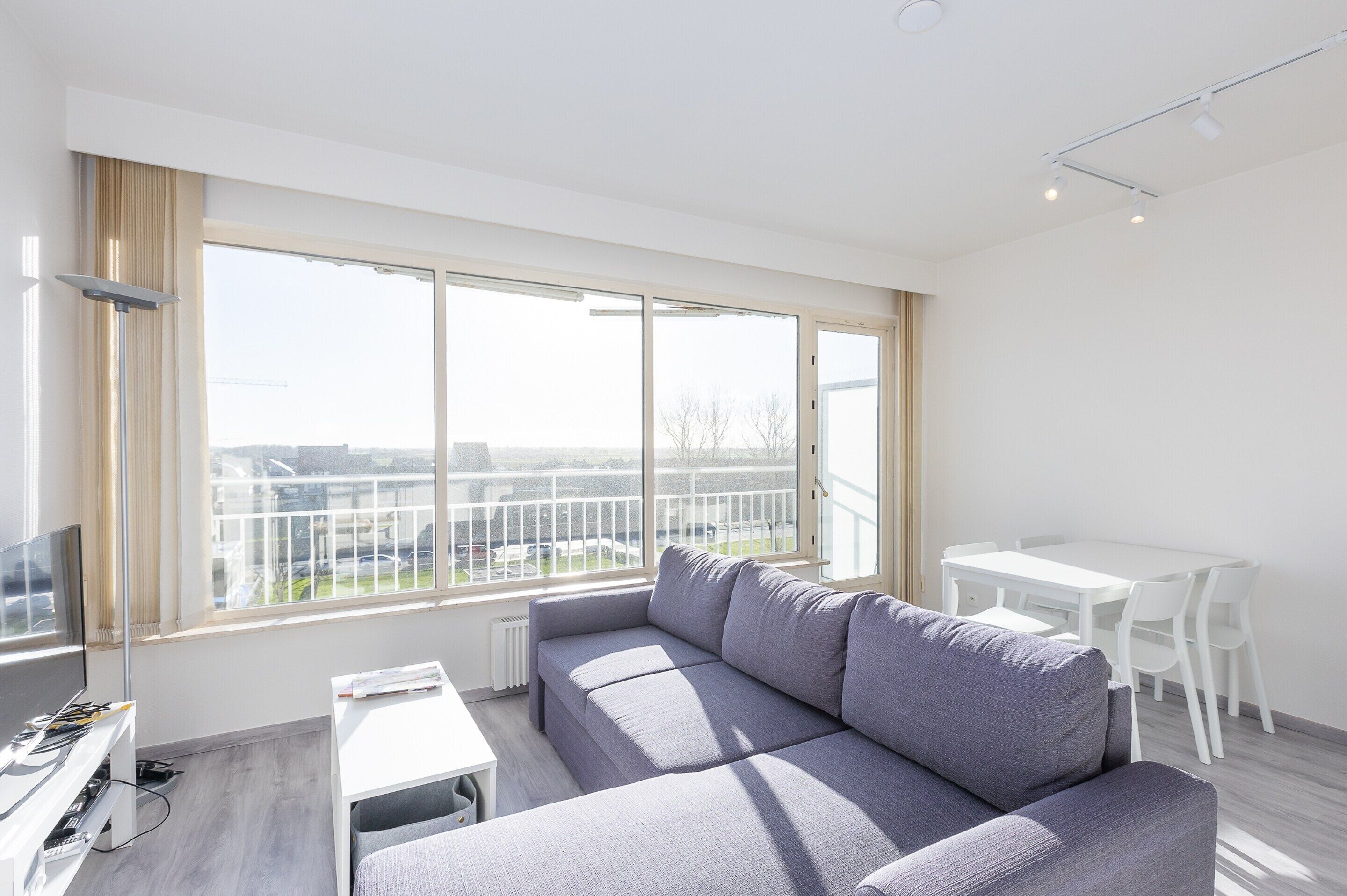 A modern living room with a grey sofa, a small white table, and a large window with a view of the city.