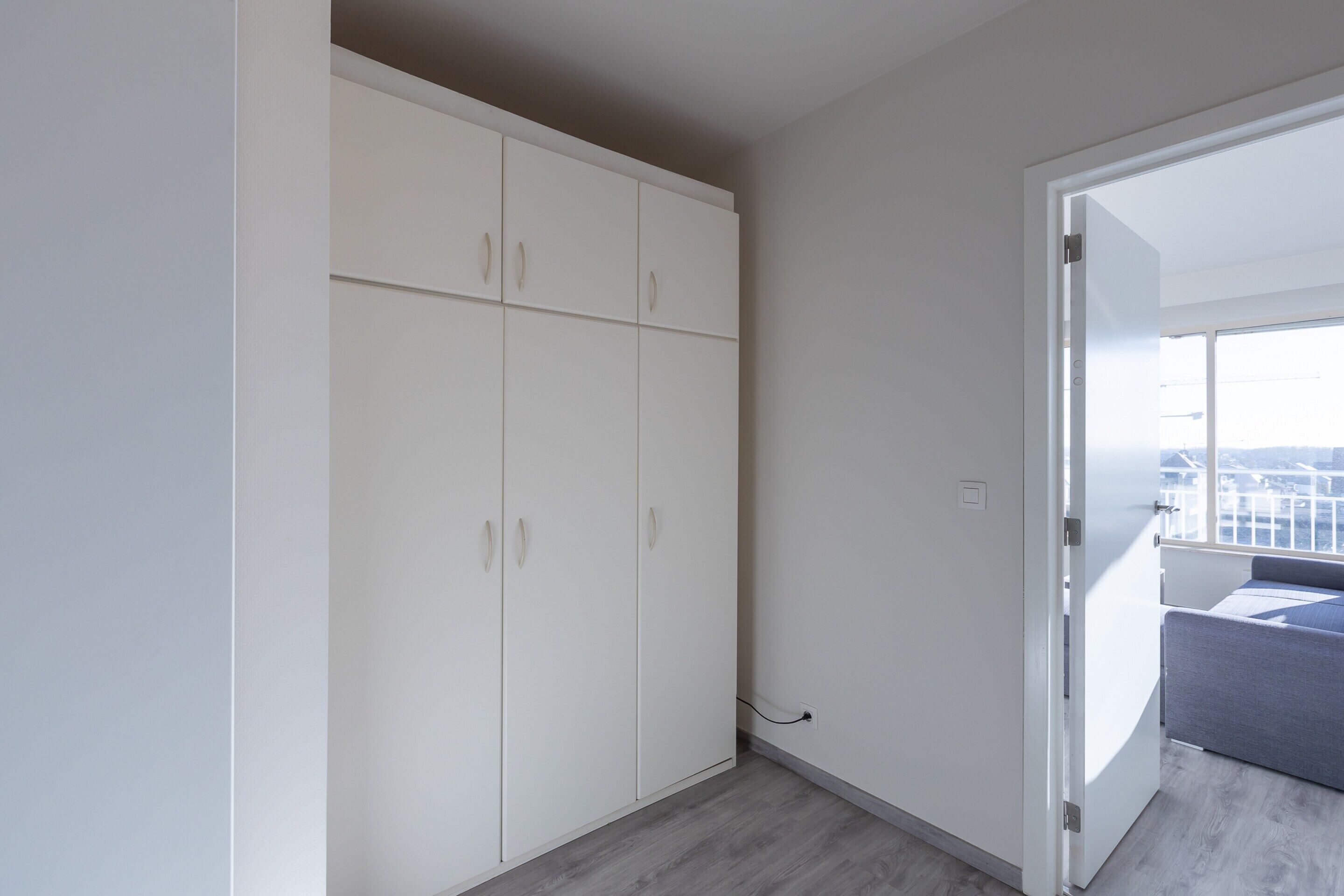 A room with a large white wardrobe, a grey sofa, and a view through an open door.