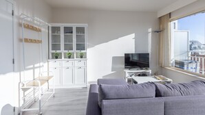 A modern living room with a grey sofa, a TV on a white stand, and a kitchen area with cabinets and a plant. - Modern, Renovated Studio With Open Views (Middelkerke)