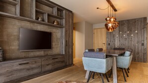 Ein modernes Wohnzimmer mit großem Flachbildfernseher an einer Holzwand, einem Essbereich mit Holztisch und blau bezogenen Stühlen sowie einem einzigartigen Kronleuchter.